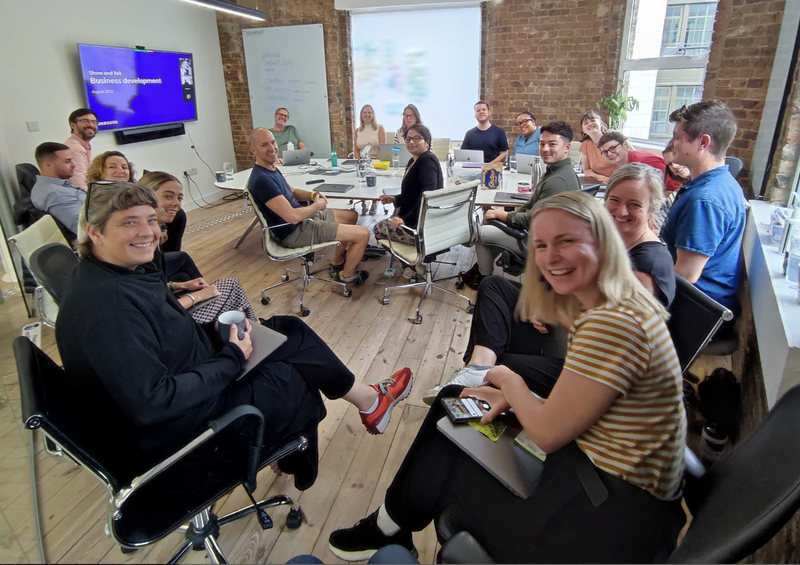 Nineteen members of the Unboxed team in a company meeting, turning to face the camera and smiling, with a presentation on a screen at the back of the room